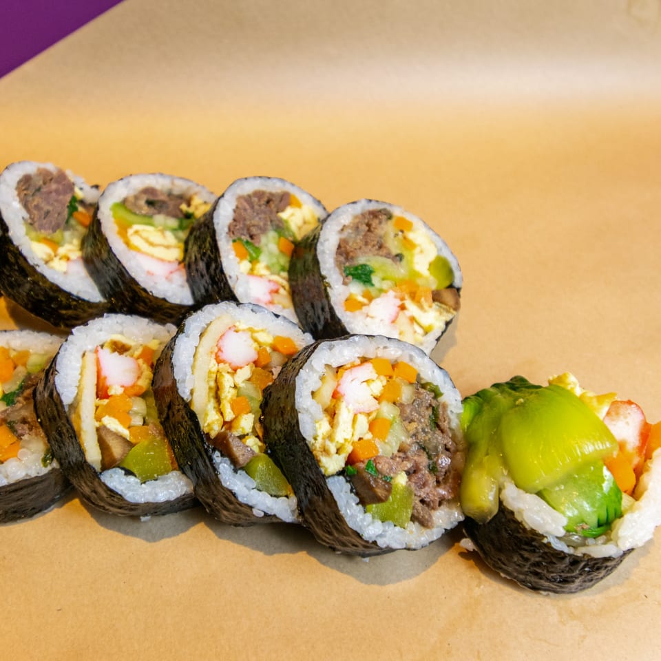 Rows of GimMeBap's Korean gimbap rolls with various fillings on a brown surface.
