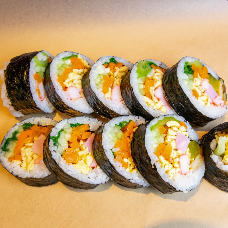 Rows of GimMeBap's Korean gimbap rolls, filled with colorful vegetables, egg, and imitation crab.